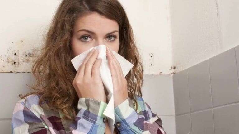 A woman is holding a cloth to her nose with both hands, and mold is visible on the wall behind her.
